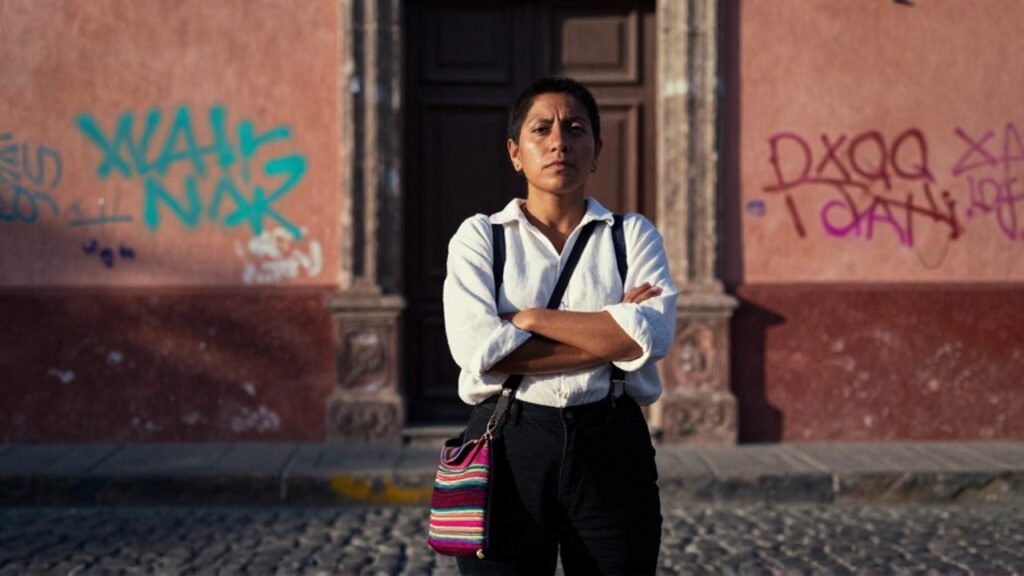 María Galindo, con el pelo muy corto y los brazos cruzados, mira desafiante a la cámara en una calle empedrada de La Paz, con una bolsa andina al hombro y un muro con grafitis de fondo.