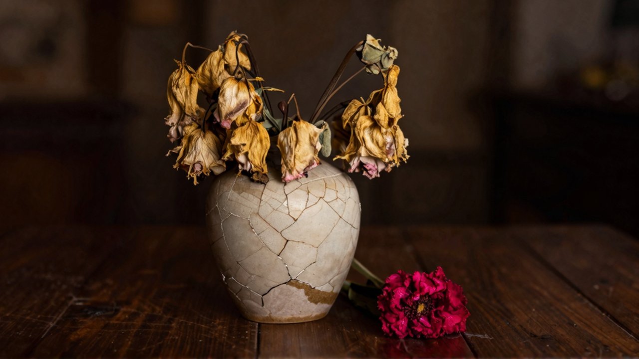 Un jarrón de cerámica agrietado con flores marchitas junto a una única flor fresca sobre una mesa de madera oscura.