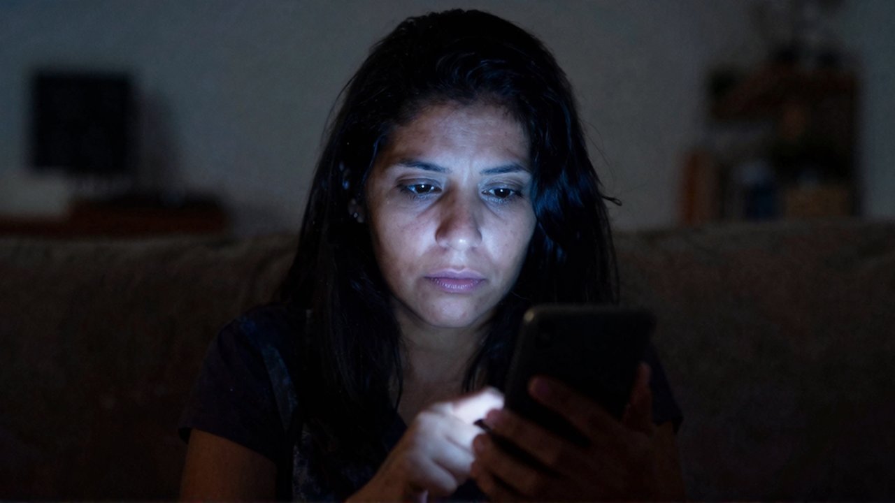 Mujer con expresión de preocupación mira la pantalla de un teléfono móvil en una habitación oscura.